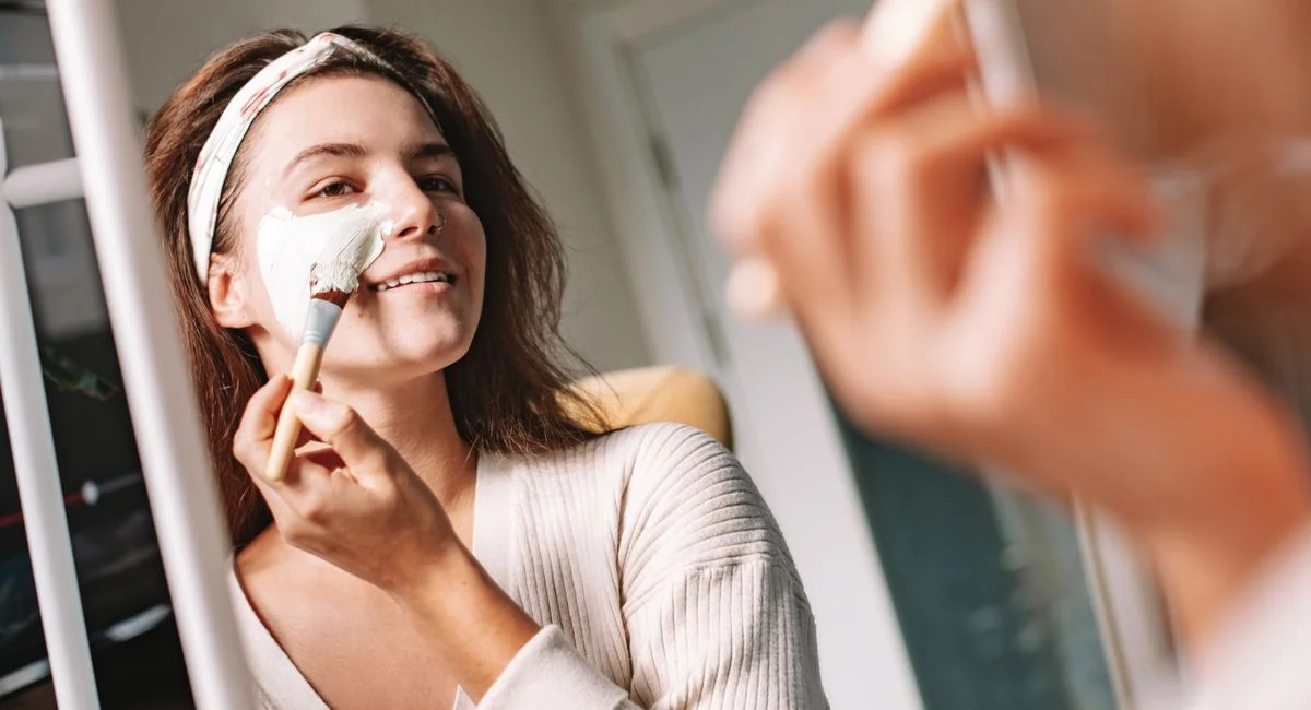 Woman enjoys a skincare routine, applying a facial mask with a brush in front of a mirror.