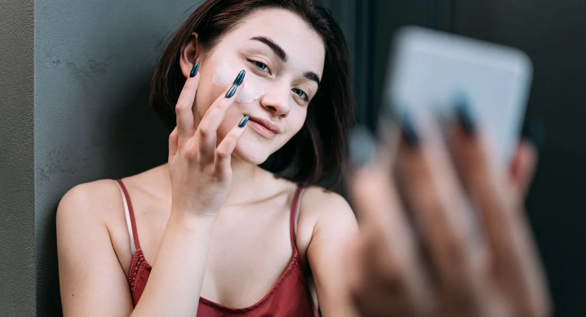 Young Caucasian woman applying moisturizer while smiling in front of a mirror indoors.