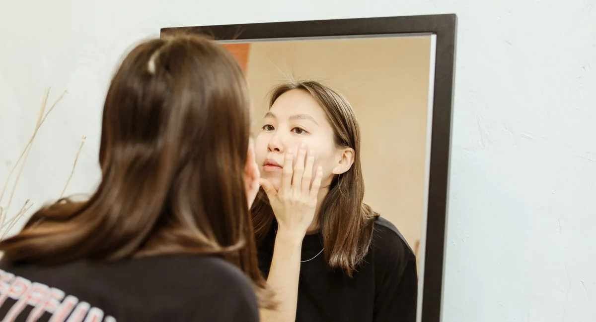 A woman in a black shirt checking her skin in front of a mirror indoors.